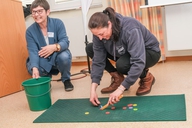 Zwei Frauen hocken auf dem Boden und spielen mit kleinen bunten Plättchen.