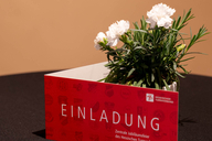 Die Einladungskarte steht vor einer Blume auf dem Tisch, sodass groß "Einladung" zu lesen ist.