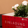 Die Einladungskarte steht vor einer Blume auf dem Tisch, sodass groß "Einladung" zu lesen ist
