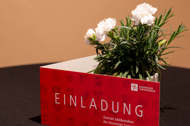 Die Einladungskarte steht vor einer Blume auf dem Tisch, sodass groß "Einladung" zu lesen ist