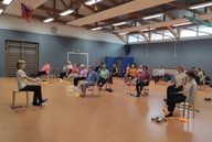 Eine Gruppe sitzt in einer Sporthalle und zeigt eine Sportübung.