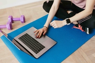 Person sitzt auf einer Sportmatte mit Laptop, daneben liegen Hanteln